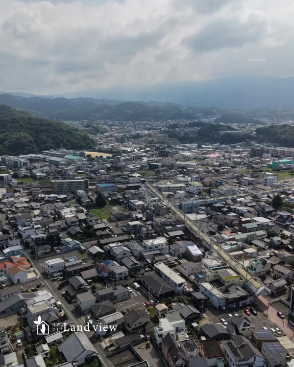 愛媛県四国中央市川之江町のJR川之江駅周辺を上空から撮影した写真。住宅密集地やアーケード、道路が広がり、遠くには緑の山並みと雲の広がる空が見える。