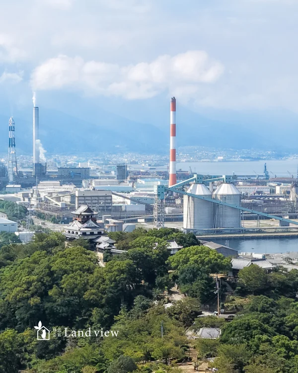 四国中央市のドローン空撮写真。緑の木々に囲まれたお城の向こうに、煙突や大型の工場設備が並ぶ工業地帯と山並みが広がる風景。